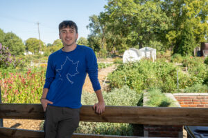 A white male student with short brown hair stands against a wooden railing in front of a garden. He wears a blue long-sleeved shirt with a white sycamore leaf on the front, and grey pants.