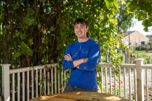 A white male student with short brown hair poses with his arms crossed in front of a white fence. He wears a blue long-sleeved shirt with a white sycamore leaf on the front. A wooden table is visible in front of him. Trees are visible in the background. 