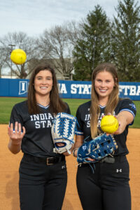 Two softball players on a softball field. On the left is a white female with dark brown hair. On the right is a white female with light brown hair. They wear black softball uniforms and hold blue and white softball mitts and softballs. The woman on the left is throwing the ball up in the air.