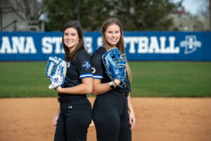 Two softball players on a softball field. On the left is a white female with dark brown hair. On the right is a white female with light brown hair. They wear black softball uniforms and hold blue and white softball mitts.