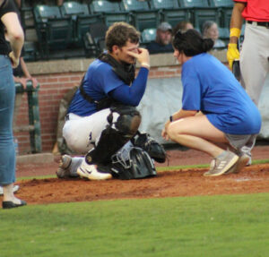 Mandy Flaig, a white woman with dark brown hair, leans down in front of a baseball catcher. They both wear blue clothing. The athlete has his right hand on his face near his eyes.
