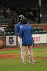 Mandy Flaig, a white woman with dark brown hair, stands on a baseball field wearing a blue T-shirt and shorts. She talks to a baseball player in a blue-and-white uniform and black helmet.