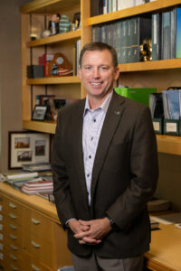 Paul Thrift, a middle-aged white male with short brown hair, stands in an office setting. He wears a dark brown suit jacket with a white undershirt. He smiles as he poses for the camera. A bookshelf with books and notebooks is visible behind him.