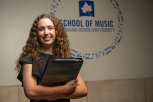 Sophia Greenwood, a white female student with long curly brown hair, poses in front of a white wall with "School of Music Indiana State University" in blue lettering on the wall. A Sycamore leaf logo is on the wall and a circular design of blue music notes is also visible. She wears a black dress and she holds a black music binder.