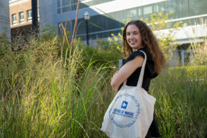 Sophia Greenwood, a white female student with long curly brown hair, poses outside with an academic building visible behind her and a small garden of green bushes. She wears a black dress. Around her left shoulder is a white tote bag with the School of Music logo printed on it.