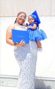 A Black female with black and red hair wears a white shimmering dress. She holds a Black infant wearing a blue dress and a blue graduation cap. The woman holds a blue diploma holder. A white wall is visible behind them.
