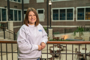 Ellie Heerema, a white female student wit shoulder-length brown hair, stands against a railing in an atrium. She wears a white long-sleeved sweatshirt with "Indiana State University Honors College" in blue lettering in the top right corner. Tables and chairs are in the atrium, along with greenery. Brick walls are visible behind her.