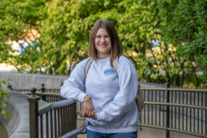 Ellie Heerema, a white female student wit shoulder-length brown hair, stands outside, posing next to a railing. She wears a white long-sleeved sweatshirt with "Indiana State University Honors College" in blue lettering in the top right corner. She also wears a backpack. Greenery is visible behind her.