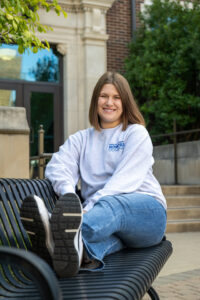 Ellie Heerema, a white female student wit shoulder-length brown hair, sits on a bench with her feet up on the bench. She wears a white long-sleeved sweatshirt with "Indiana State University Honors College" in blue lettering in the top right corner. A brick building is visible behind her. Greenery is also visible behind her.