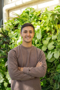Spencer Limcaco, a male student with short brown hair, stands in front of green plants. He wears a brown long-sleeved shirt and his arms are crossed.
