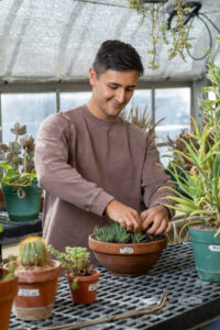 Spencer Limcaco, a male student with short brown hair, stands in a greenhouse wearing a brown long-sleeved shirt. He is working on a potted green plant. Other potted plants are visible around him.