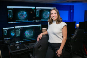 Payton Ferency, a white female student with shoulder-length brown hair, stands in a computer lab with computer monitors visible behind her. She wears a white short-sleeve sweater and black dress pants.