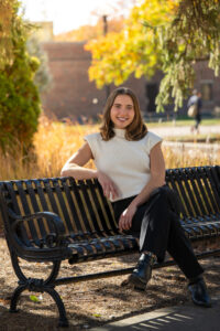 Payton Ferency, a white female student with shoulder-length brown hair, sits with her legs crossed on a black bench outside. She wears a white short-sleeve sweater and black dress pants. Fall foliage is visible behind her.