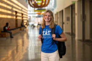 Peyton Heagy, a white female student with blonde hair, poses in a hallway with a black backpack around her shoulders. She wears white pants and a blue T-shirt with STATE Honors College in white lettering.