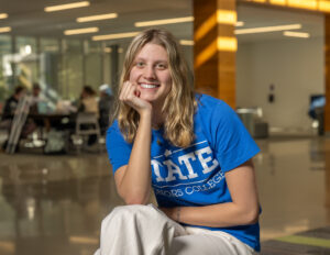 Peyton Heagy, a white female student with blonde hair, sits in a hallway with her chin resting on the palm of her right hand. She wears white pants and a blue T-shirt with STATE Honors College in white lettering. Other students are blurred in the background.
