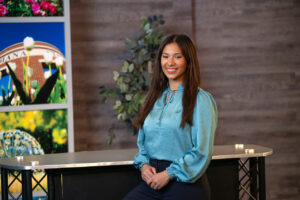 Piper Watkins, a female student with tanned skin and long brown hair, poses in front of a table in a media room. She wears a teal shirt. A plant is visible behind her. To the left of the table are photos of Indiana State's campus.