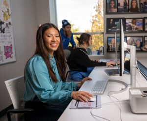 Piper Watkins, a female student with tanned skin and long brown hair, sits in a media lab. She wears a teal shirt. A computer monitor is on a table in front of her. Two students are visible behind her.