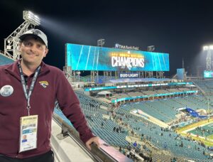 A white male wearing a maroon long-sleeved jacket with a Jacksonville Jaguars logo in the top right corner, a baseball cap, and a lanyard. He poses at the top of a football stadium. A digital sign reads: "AFC South Division Champions 2022." 