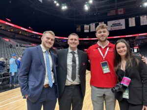 Four white individuals pose on a basketball court in a basketball arena. 