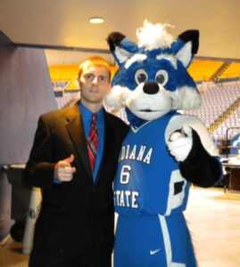 A white male wearing a black dress suit and a blue undershirt poses next to Sycamore Sam, a blue-and-white fox-like mascot wearing an Indiana State jersey. They stand in a hallway leading to a basketball arena. 