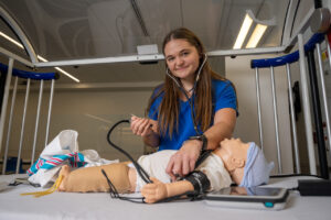 A white female student with long brown hair stands in a healthcare simulation lab. She wears blue nursing scrubs with Indiana State University Nursing in white lettering in the top right corner. She also wears a stethoscope. She is attending to a simulated baby dummy in a hospital crib. 