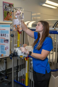 A white female student with long brown hair stands in a healthcare simulation lab. She wears blue nursing scrubs with Indiana State University Nursing in white lettering in the top right corner. An IV machine is visible in front of her. A hospital crib with a simulation baby dummy is visible behind her.