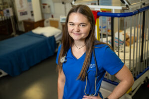 A white female student with long brown hair stands in a healthcare simulation lab. She wears blue nursing scrubs with Indiana State University Nursing in white lettering in the top right corner. She also wears a stethoscope. A hospital crib with a simulation baby dummy is visible behind her. 