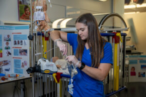 A white female student with long brown hair stands in a healthcare simulation lab. She wears blue nursing scrubs with Indiana State University Nursing in white lettering in the top right corner. An IV machine is visible in front of her. A hospital crib with a simulation baby dummy is visible behind her. 