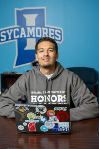 A tanned male student with short black hair sits at a desk in an office, wearing a grey sweatshirt with Indiana State University Honors College in white lettering. A blue wall is visible in the background with the Sycamores logo printed on the wall. A laptop covered in stickers is visible on the desk in front of the student.