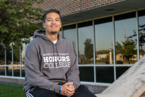 A tanned male student with short black hair wears a grey sweatshirt with Indiana State University Honors College in white lettering. He sits outside with a building visible in the background, with windows and a brick wall.