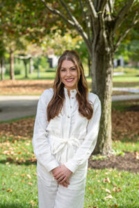 A white female with long wavy brown hair posing outside wearing a white outfit. She stands in front of a tree. Leaves are visible in the background.