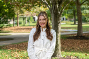 A white female with long wavy brown hair posing outside wearing a white outfit. She stands in front of a tree. Leaves are visible in the background.