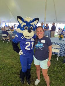 Nicole Otte, a white woman with blond hair, stands next to Sycamore Sam, Indiana State University's mascot, in a tent. She wears a dark grey T-shirt with a blue sycamore leaf on the front, and light blue shorts. Other people are visible behind them. 