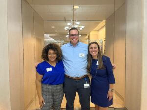 Nicole Otte (right), a white woman with dark blond hair, wears a blue dress. To her left is a white male with short brown hair and glasses. He wears a blue dress shirt and dark grey dress pants. On the far left is a Black woman with shoulder-length curly brown hair. She wears a blue shirt and grey checkered dress pants. Glass doors and a hallway are visible behind them. 