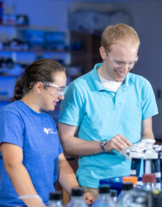 In a chemistry lab, Justin Miller (right), a white male with short blond hair, stands next to a student with a brown braid wearing a blue shirt and safety glasses. They look down at the lab table while Miller holds an instrument in his left hand. Lab equipment are on shelves behind them, and are visible on a table in front of them. There is a blue tint to the photo.