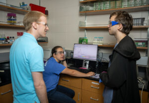 In a chemistry lab, Justin Miller (left), a white male with short blond hair, stands with two students. One wears a blue T-shirt and sits on a chair in front of a computer, and the other is standing on the right wearing a black jacket. They each wear protective glasses. A graph is visible on the computer screen. Shelves with laboratory equipment are visible on the walls opposite them. 