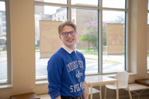 Craig Robinson, a white young man with short brown hair, poses in a row of glass windows. He appears to be in a lobby area of a building. He wears glasses, a blue sweatshirt and black pants. The sweatshirt has the Indiana State logo and white lettering: Indiana State Sycamores. A white table and white chairs are visible behind him. 