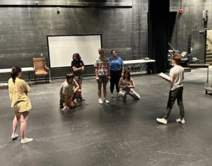 A diverse group of individuals on a theater stage, wearing a variety of clothing. The person on the far right holds a binder with a script. A whiteboard is visible in the background, along with a brown chair. 