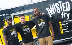 Three diverse men stand in front of a black-and-yellow food truck with white lettering Twisted Fry on the front. They wear matching black T-shirts with the Twisted Fry logo on the front.