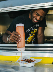 Devyn Mikell, a young Black male, stands inside a food truck wearing a black T-shirt. He holds a squirt bottle with an orange-color sauce. He pours the sauce on a basket of food. 