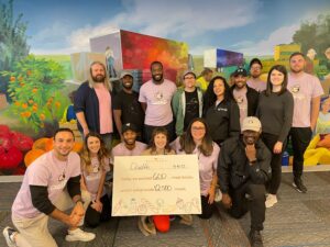 A diverse group of 15 individuals posing in front of a colorful wall mural. They hold up a white sign: "Qualifi, 4-14-22, Today we packed 600 meal boxes, which will provide 12,500 meals." 