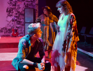 A theater production from "Hamlet." On the left is a white male with blonde hair. He wears a denim jacket, dark pants, and a brown bandana around his forehead. On the right is a white female with shoulder-length curly brown hair. She wears a white dress with a floral robe. Another woman is visible behind them. The set looks like a rock concert with a red guitar visible in the background. 