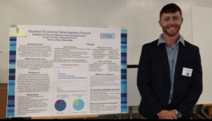 Jackson Taylor, a white male with short red hair, poses next to a presentation board with "Marshall Economic Development Project" in blue lettering near the top. He wears a black dress suit with a light blue dress shirt underneath.
