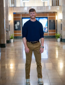 Jackson Taylor, a white male with short red hair, poses inside an academic building wearing a blue long-sleeved sweater and khaki pants. He stands in the middle of a hallway with digital displays visible behind him. Glass light fixtures hang from the ceiling.