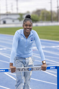 A Black female student with black hair tied into a tight bun. She wears a light blue athletic shirt and blue jeans. She stands on a blue outdoor running track. She leans against a blue hurdle for track events. "Indiana State University" and the athletic logo is on the hurdle.