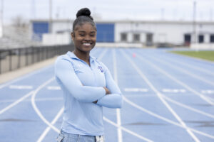 A Black female student with black hair tied into a tight bun. She wears a light blue athletic shirt, Her arms are crossed over her chest. She stands on a blue outdoor running track.