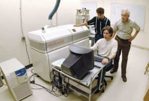 Three individuals in a laboratory setting, and they are gathered around a large scientific instrument. One person is seated at a computer workstation while the other two stand nearby observing. A small unit is visible on the left, and a whiteboard with writing is on the wall in the background. 
