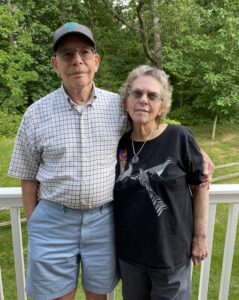 Two older individuals stand closely together on a porch with greenery visible behind them. The man on the left wears a checkered shirt, light blue shorts, and a baseball cap. The woman on the right wears a black T-shirt with an abstract bird design and dark pants. 