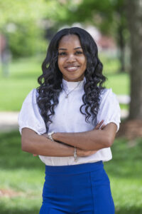 A young Black female with long, wavy black hair stands outdoors with her arms crossed. She is wearing a white blouse with puffed sleeves and blue pants. The background shows green grass and trees.