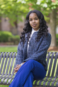 A young Black female with long, wavy black hair sits on a park bench. She is wearing a blue tweed jacket over a white shirt and blue pants. The background shows green trees and bushes, indicating an outdoor setting.